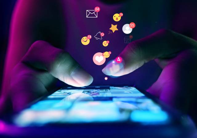 Manos utilizando un teléfono inteligente mientras aparecen iconos de notificaciones e interacciones en pantalla, representando estrategias de Social Media Search Optimization para aumentar la visibilidad en redes sociales.