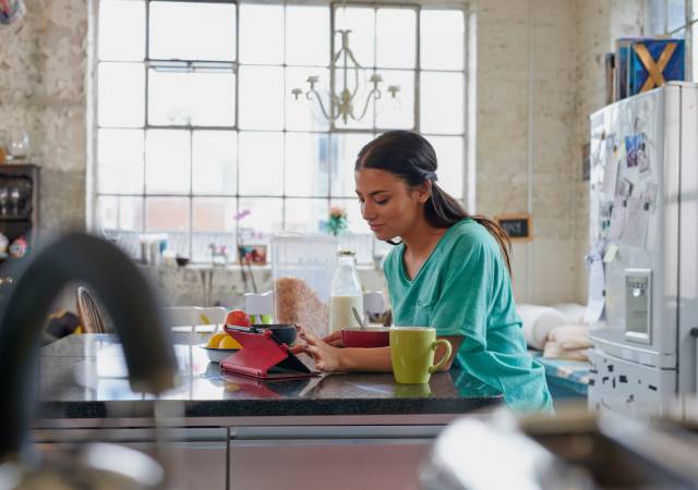 Mujer utilizando una tablet en la cocina de su casa, junto a una taza y varios alimentos sobre la encimera