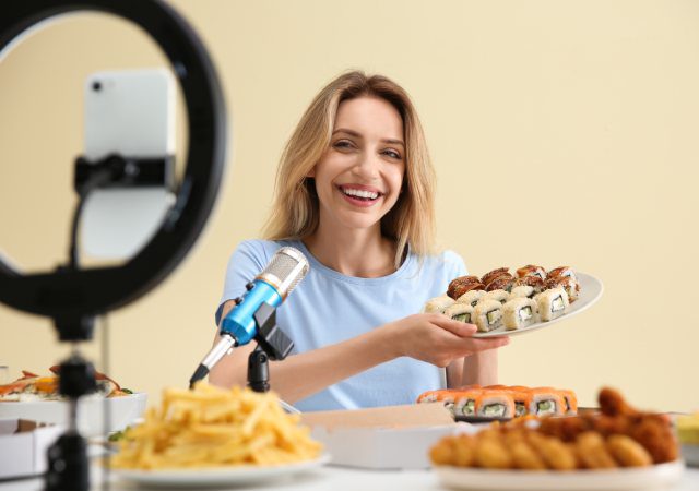 Mujer grabando contenido gastronómico con un móvil, un aro de luz y un micrófono mientras muestra un plato de sushi